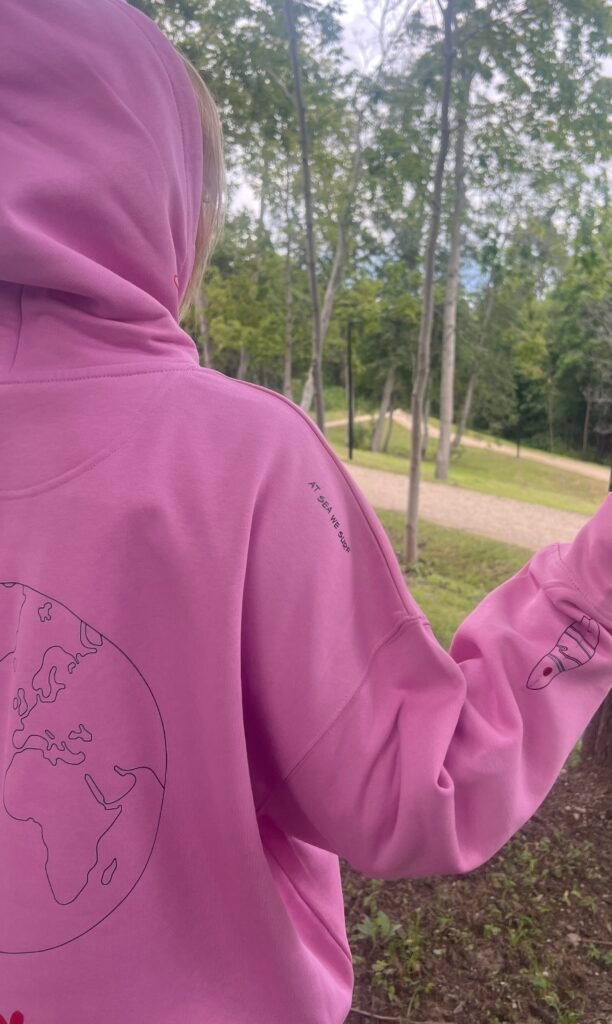 A pink Sun Sets West hoodie worn beside a tree